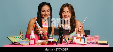 Zwei glückliche weibliche Influencer untersuchen Kosmetikprodukte, während sie online vor blauem Hintergrund übertragen Stockfoto