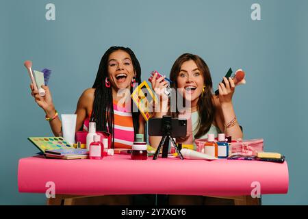 Zwei glückliche weibliche Influencer halten verschiedene Schönheitsprodukte, während sie Make-up Tutorial auf blauem Hintergrund aufzeichnen Stockfoto