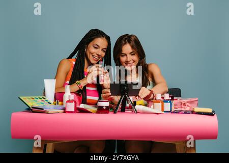 Zwei glückliche junge Frau, die Make-up-Tutorial aufnimmt, während sie am Schreibtisch vor blauem Hintergrund sitzt Stockfoto