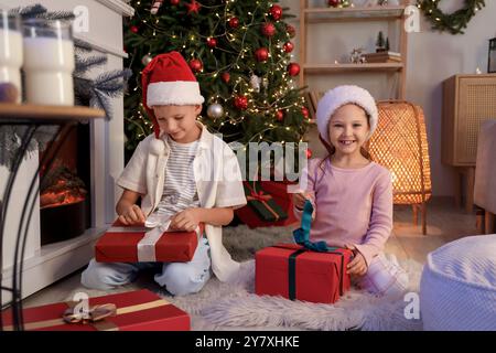 Niedliche kleine Kinder mit Weihnachtsmützen, die Geschenke zu Hause am Weihnachtsabend auspacken Stockfoto