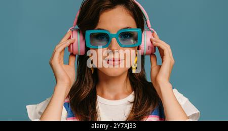 Porträt einer schönen jungen Frau in Kopfhörern und trendigen Brillen, die Musik auf blauem Hintergrund genießen Stockfoto