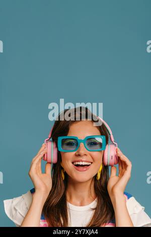Porträt einer glücklichen jungen Frau in Kopfhörern und trendigen Brillen, die Musik auf blauem Hintergrund genießen Stockfoto