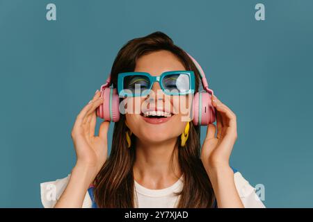 Porträt einer fröhlichen jungen Frau in Kopfhörern und trendigen Brillen, die Musik auf blauem Hintergrund genießen Stockfoto