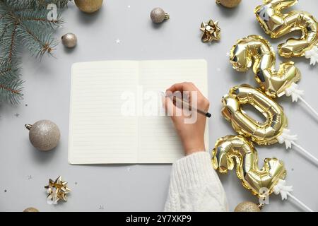 Frau schreibt Pläne für 2025 Jahre in Notizbuch und Weihnachtskugeln auf hellem Hintergrund Stockfoto