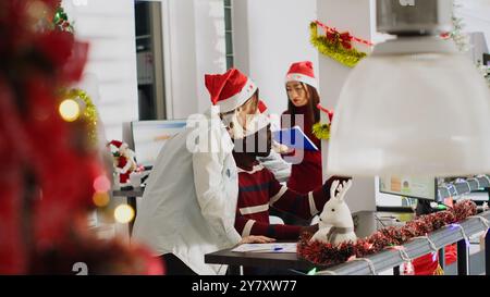 afroamerikanischer Angestellter, der im weihnachtlich verzierten Büro um Hilfe bittet. Firmenmitarbeiter, die sich Finanzdaten auf Weihnachten ansehen, schmücken den Computerbildschirm am Arbeitsplatz Stockfoto