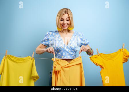 Glückliche junge Frau, die ihre bunten Kleider auf einer Wäscheleine vor blauem Hintergrund hängt Stockfoto