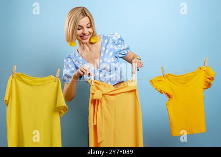 Schöne junge Frau, die ihre bunten Kleider auf Kleiderleinen vor blauem Hintergrund hängt Stockfoto