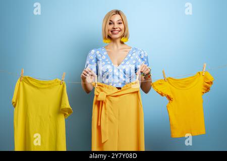 Glückliche junge Frau, die ihre bunten Kleider auf einer Wäscheleine vor blauem Hintergrund hängt Stockfoto