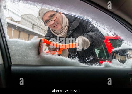 Ein reifer Mann reinigt sein Auto an einem frostigen Wintertag vom Schnee. Ein Mann kratzt an einem kalten Wintermorgen an der Frontscheibe seines Autos, Innenansicht Stockfoto