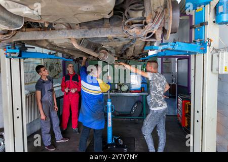 Erfahrener Senior-Lehrer, der unter dem Auto auf der hydraulischen Rampe steht und seinen Schülern an der Berufsschule den Automobilhandel unterrichtete Stockfoto