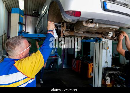 Reifer erfahrener Lehrer, der unter dem Auto auf der hydraulischen Rampe steht und seinen Schülern in der Berufsschule den Automobilhandel unterrichtete Stockfoto