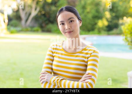 Draußen, lächelnde junge asiatische Frau in gelb gestreiftem Hemd mit überkreuzten Armen im Garten Stockfoto