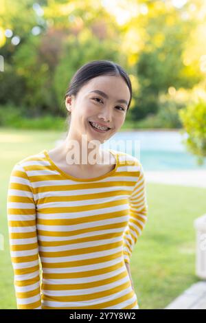 Lächelnde junge asiatische Frau in gelb gestreiftem Hemd, die draußen im Garten steht Stockfoto