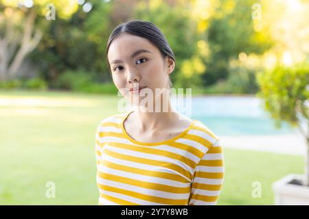 Lächelnde junge asiatische Frau in gelb gestreiftem Hemd, die draußen im Garten steht Stockfoto