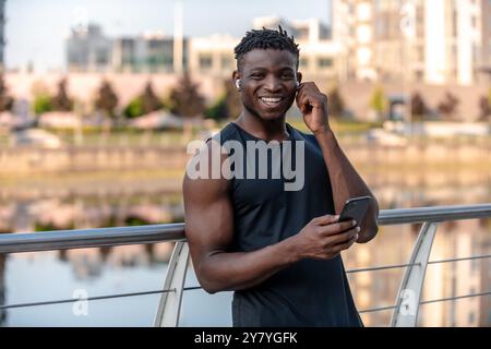 Glücklicher afrikanischer Sportler, der das Telefon benutzt und Kopfhörer anpasst, während er sich nach dem Training im Freien ausruht Stockfoto