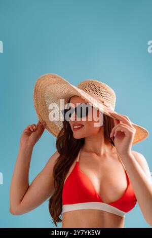 Attraktive junge Frau im Bikini mit elegantem Hut vor hellblauem Hintergrund Stockfoto