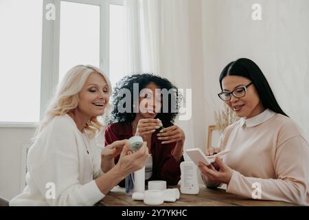 Drei glückliche, reife Frauen diskutieren über Schönheitsbehandlungen, während sie zusammen am Schreibtisch sitzen Stockfoto