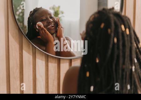 Glückliche afrikanische Frau in Plus Größe, die das Gesicht mit Wattepad reinigt und auf den Spiegel im Badezimmer blickt Stockfoto