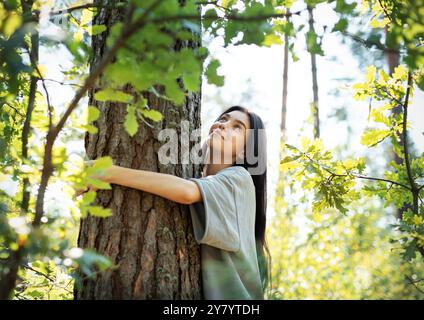 Eine Frau steht in einem lebendigen Wald und umhüllt liebevoll einen Baum. Das Sonnenlicht durchströmt die Blätter und verleiht der ruhigen Atmosphäre Wärme. Stockfoto