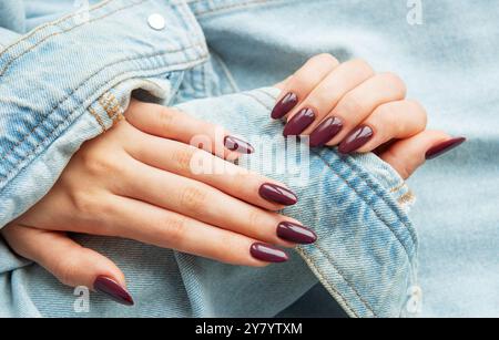 Zwei Hände mit wunderschönen burgunderroten Nägeln, die sanft auf eine lässige Jeansjacke gelegt werden. Das Ambiente unterstreicht einen schicken und trendigen Stil mit einer entspannten Atmosphäre Stockfoto