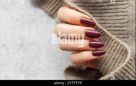 Eine gut gepflegte Hand zeigt dunkle, burgunderrote Nägel und hält einen gemütlichen Strickpullover. Die warmen Töne sorgen für ein einladendes, modisches Ambiente Stockfoto