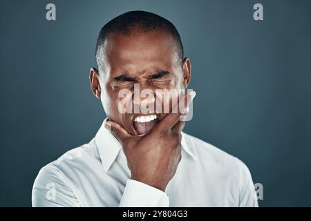 Erstellen eines Gesichts. Unzufriedener junger afrikanischer Mann im Hemd, der die Augen geschlossen hält und vor grauem Hintergrund brüllt Stockfoto