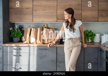 Junge Frau neben Papiertüten voller Nahrung in der Küche. Lebensmittel, die online bestellt und per Kurier geliefert werden Stockfoto