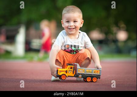 Ein fröhlicher kleiner Junge hockt sich auf dem Boden und lächelt, während er mit einem bunten Spielzeugwagen auf einem Spielplatz voller Kinder und Grün spielt. Stockfoto