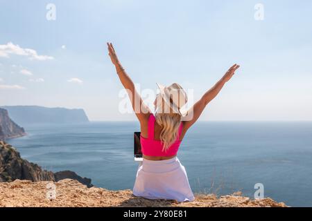 Eine Frau sitzt auf einem Hügel mit Blick auf das Meer mit einem Laptop im Schoß. Sie trägt ein rosafarbenes Oberteil und einen Strohhut. Konzept der Entspannung und Stockfoto