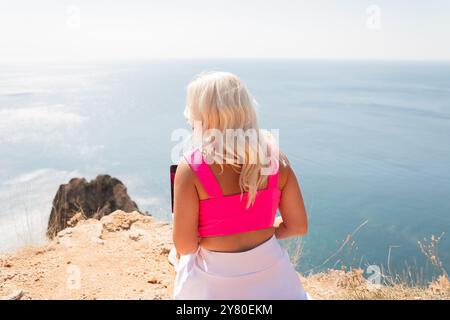 Eine Frau in einem pinkfarbenen Tanktop sitzt auf einer felsigen Klippe mit Blick auf den Ozean. Sie benutzt einen Laptop, arbeitet oder durchsucht den Stockfoto