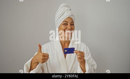 Hispanische Frau lächelt in Bademantel und Handtuch Turban, zeigt die Daumen hoch und zeigt eine Kreditkarte auf weißem Hintergrund. Stockfoto