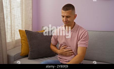 Junger Mann, der drinnen auf einer Wohnzimmercouch sitzt und seinen Magen unbehaglich hält, ein rosafarbenes Hemd trägt, mit Kissen und Vorhängen im Hintergrund Stockfoto