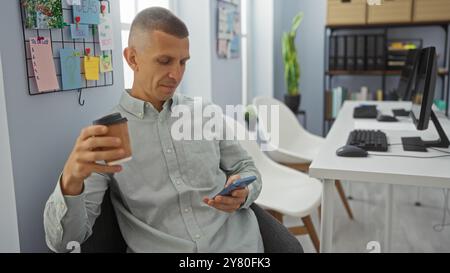 Junger Mann im Büro, der Kaffeetasse und Smartphone hält, umgeben von Büromaterialien und Dekorationen an der Wand Stockfoto