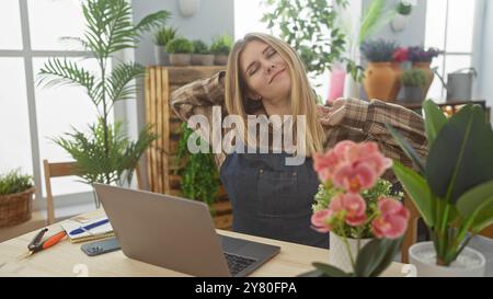 Eine junge blonde Frau dehnt sich zufrieden in ihrem von Pflanzen umgebenen Blumenladen und zeigt einen entspannten Unternehmer. Stockfoto