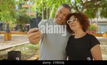 Ein Paar mittleren Alters macht gemeinsam ein Selfie in einem urbanen Outdoor-Park, um einen Moment der Liebe und Freude festzuhalten. Stockfoto