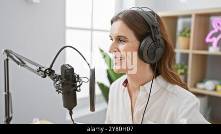 Eine Frau mittleren Alters mit Headset spricht in einem Podcast-Studio in ein Mikrofon und strahlt Professionalität und Positivität aus. Stockfoto