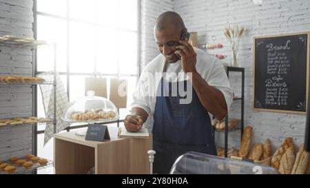 Junger Mann, der telefoniert, während er Notizen in einer gemütlichen Bäckerei mit Regalen voller Backwaren und Brot in einem Innengeschäft schreibt Stockfoto