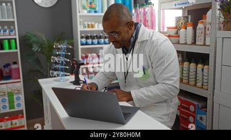 Junger Mann, der in einer Apotheke arbeitet, einen Labormantel und eine Brille trägt, Notizen schreibt, während er einen Laptop benutzt, umgeben von Medikamenten und Produkten. Stockfoto