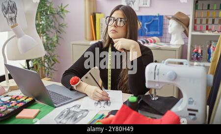 Nachdenkliche Frau skizziert ein Kleid in einem geschäftigen Atelier mit Nähaccessoires und einem Laptop. Stockfoto