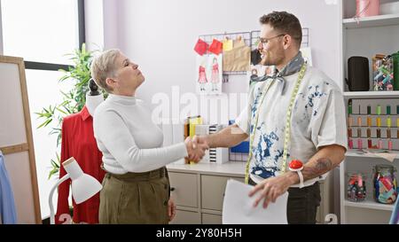 Ein Mann und eine Schneiderin schüttelt in ihrem Atelier die Hand, umgeben von Modeskizzen und bunten Fäden. Stockfoto