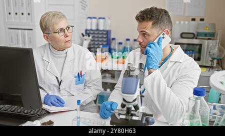 Eine Wissenschaftlerin und ihr männlicher Kollege in Labormänteln analysieren Proben in einem Labor, während der Mann am Telefon spricht. Stockfoto
