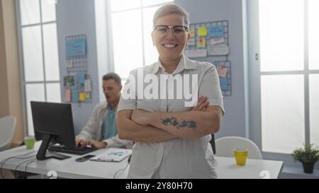 Frau lächelt selbstbewusst mit gekreuzten Armen in einem Büro, während ein Mann im Hintergrund am Computer arbeitet, was auf eine kollaborative Geschäftsumgebung hinweist Stockfoto