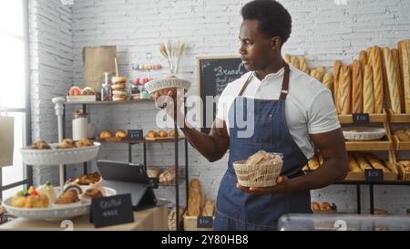 Junger Mann in einer Bäckerei, der einen Korb mit Gebäck hält, umgeben von Backwaren und Regalen, gefüllt mit Brotlaiben. Stockfoto