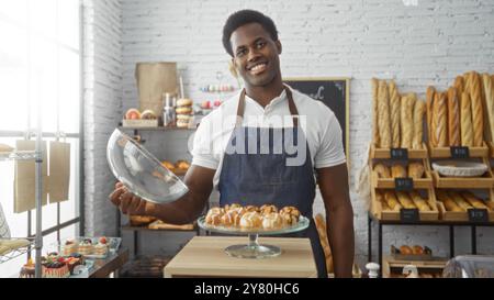 Junger Mann, der in der Bäckerei lächelt und im Hintergrund Backwaren mit Brotlaiben präsentiert Stockfoto