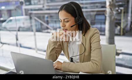 Eine professionelle junge Frau mit Headset konzentriert sich in einem hellen Büro auf ihren Laptop. Stockfoto