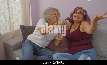 Zwei Frauen mittleren Alters singen zusammen mit einer Fernbedienung als Mikrofon in einem gemütlichen Wohnzimmer und zeigen ihre fröhliche Freundschaft und ihre lustige Bindung an Mutter Stockfoto