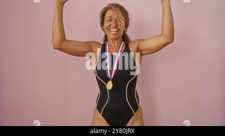 Frau mittleren Alters in schwarzem Badeanzug mit Goldmedaille lächelnd und triumphierend die Arme vor rosa Hintergrund Stockfoto