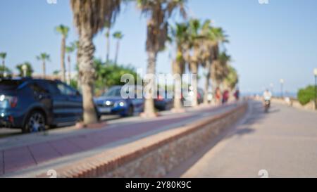 Unscharfer Blick auf eine von Palmen gesäumte Strandpromenade, geparkte Autos und Leute, die spazieren gehen, einschließlich eines Mannes, der an einem sonnigen Tag in der Nähe des Strandes radelt Stockfoto