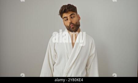 Junger hispanischer Mann mit einem Bart, der sich humorvoll in einem weißen Bademantel vor weißem Hintergrund posiert Stockfoto