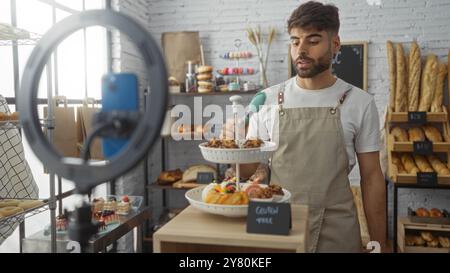 Junger Mann, der in der Bäckerei ein Video mit Smartphone aufnimmt, in dem Gebäck und Brot gezeigt werden, während er eine Schürze trägt Stockfoto